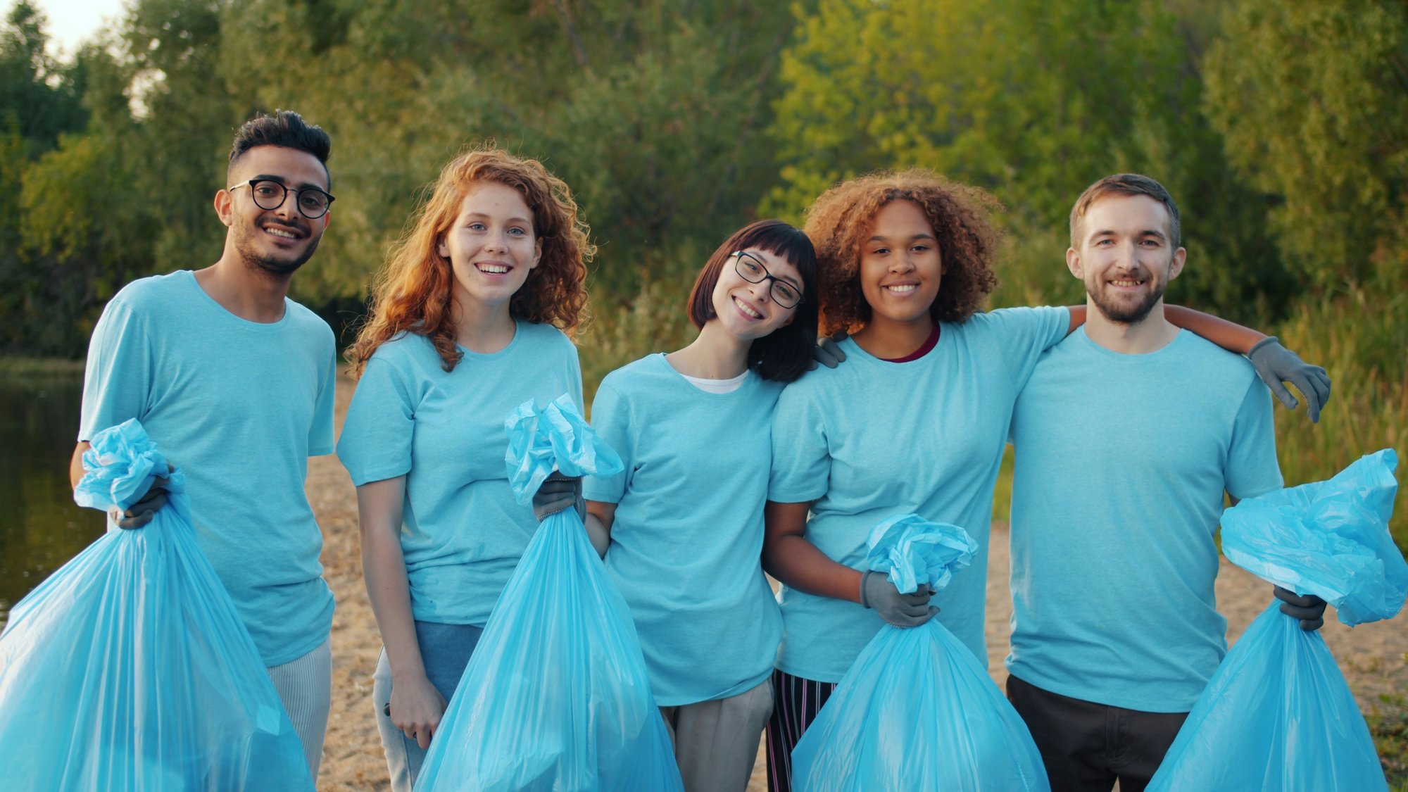 A picture of 5 cleaning staff smiling, wearing blue t shirts and holding blue garbage bags.